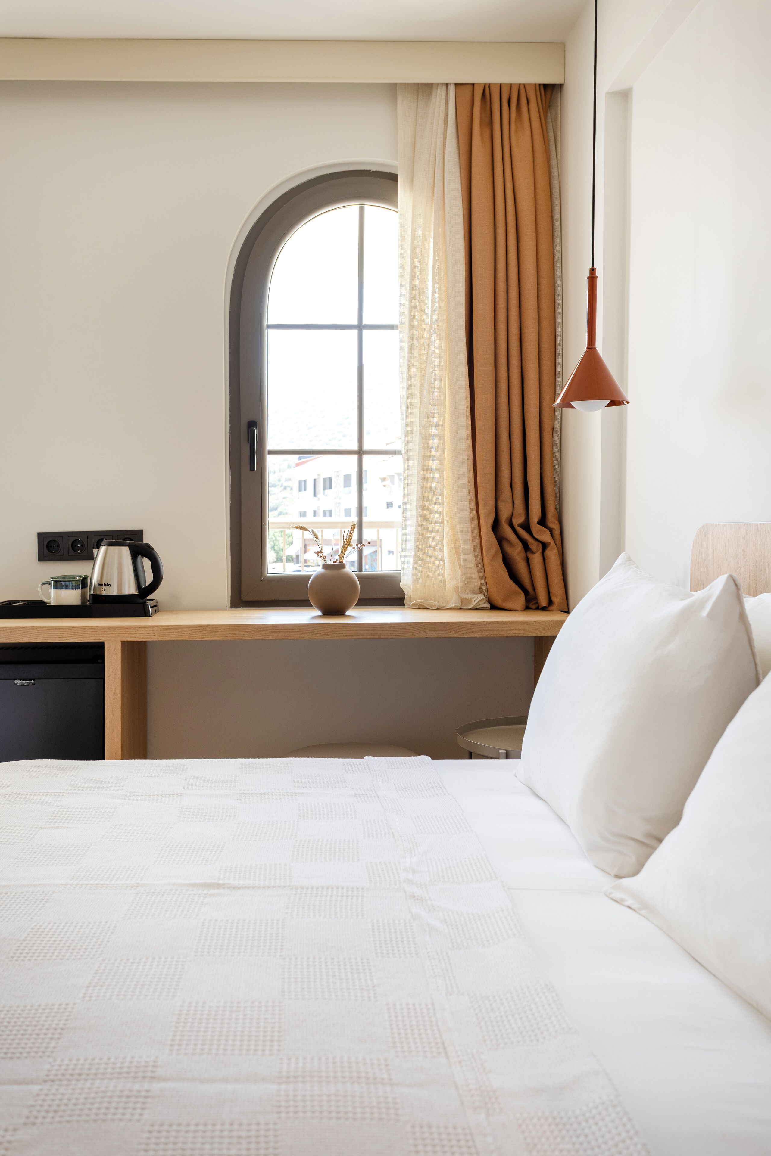 A hotel room with a large bed, a window with curtains, a desk with a kettle, and a vase with dried flowers.