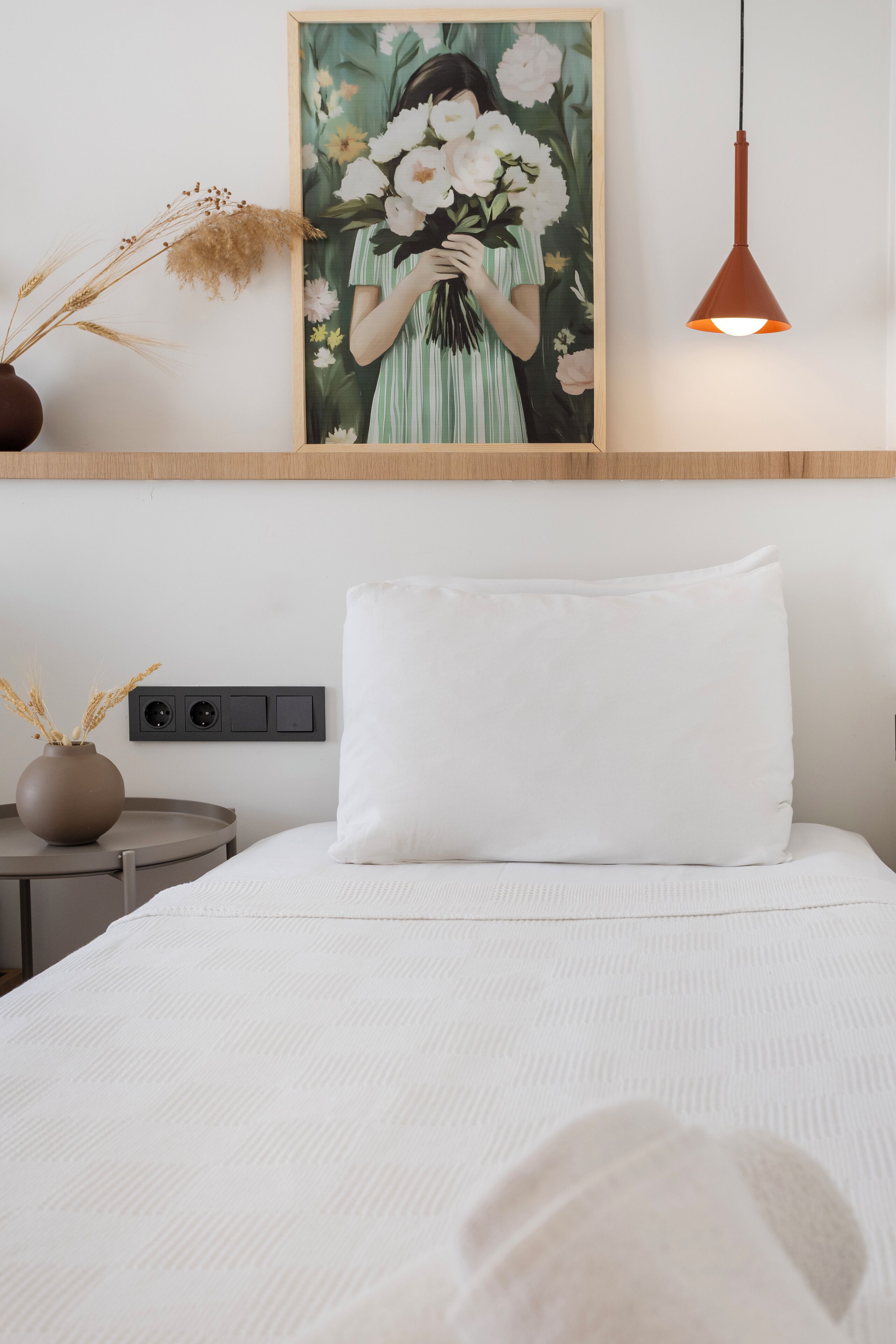 Ein ordentlich eingerichtetes Schlafzimmer mit einem Bett, einem Nachttisch mit einer Vase, einer Wandleuchte und einem gerahmten Blumenbild.