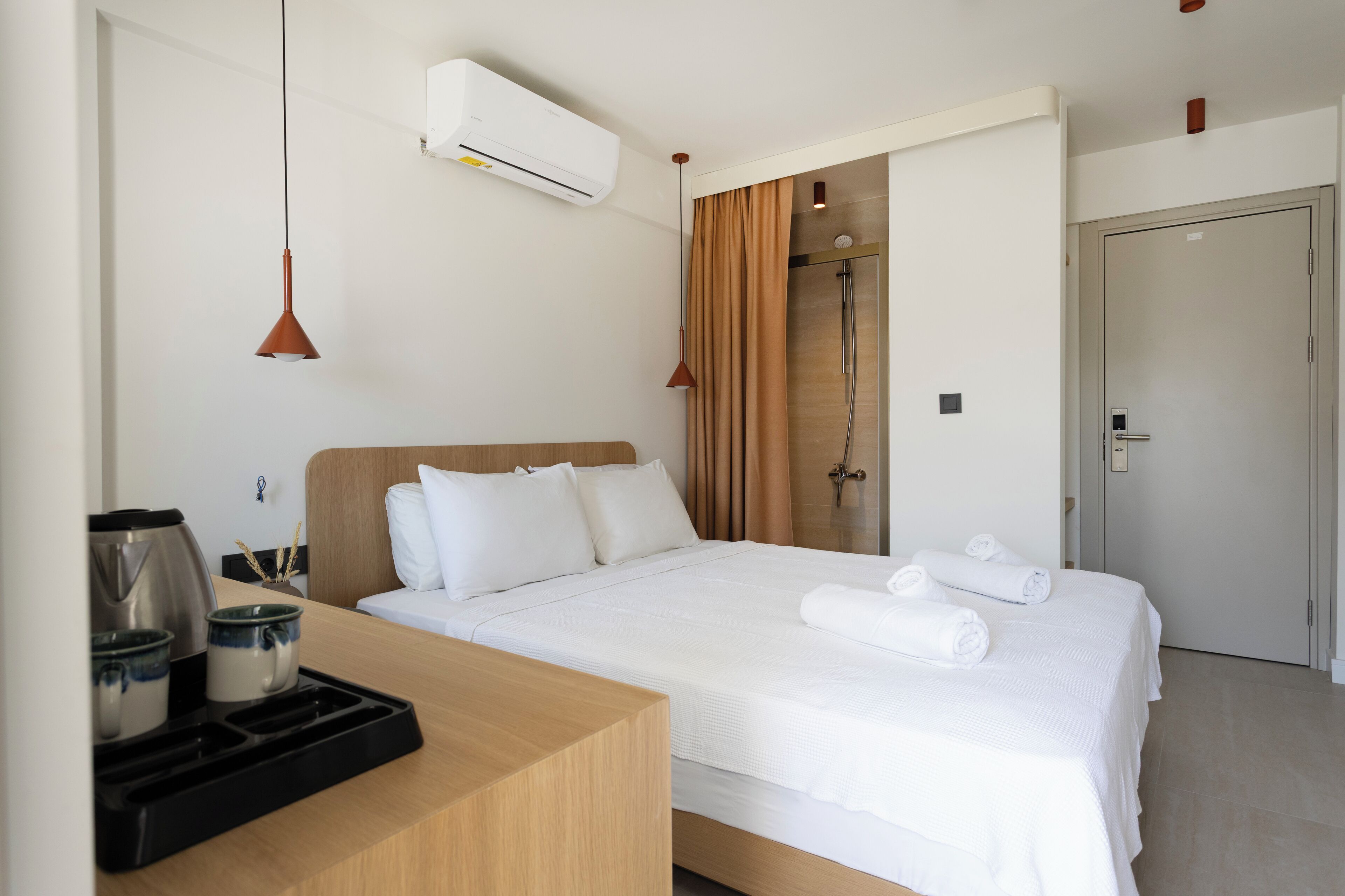 A hotel room with a bed, a bedside table with a kettle and cups, a shower area, and a door.