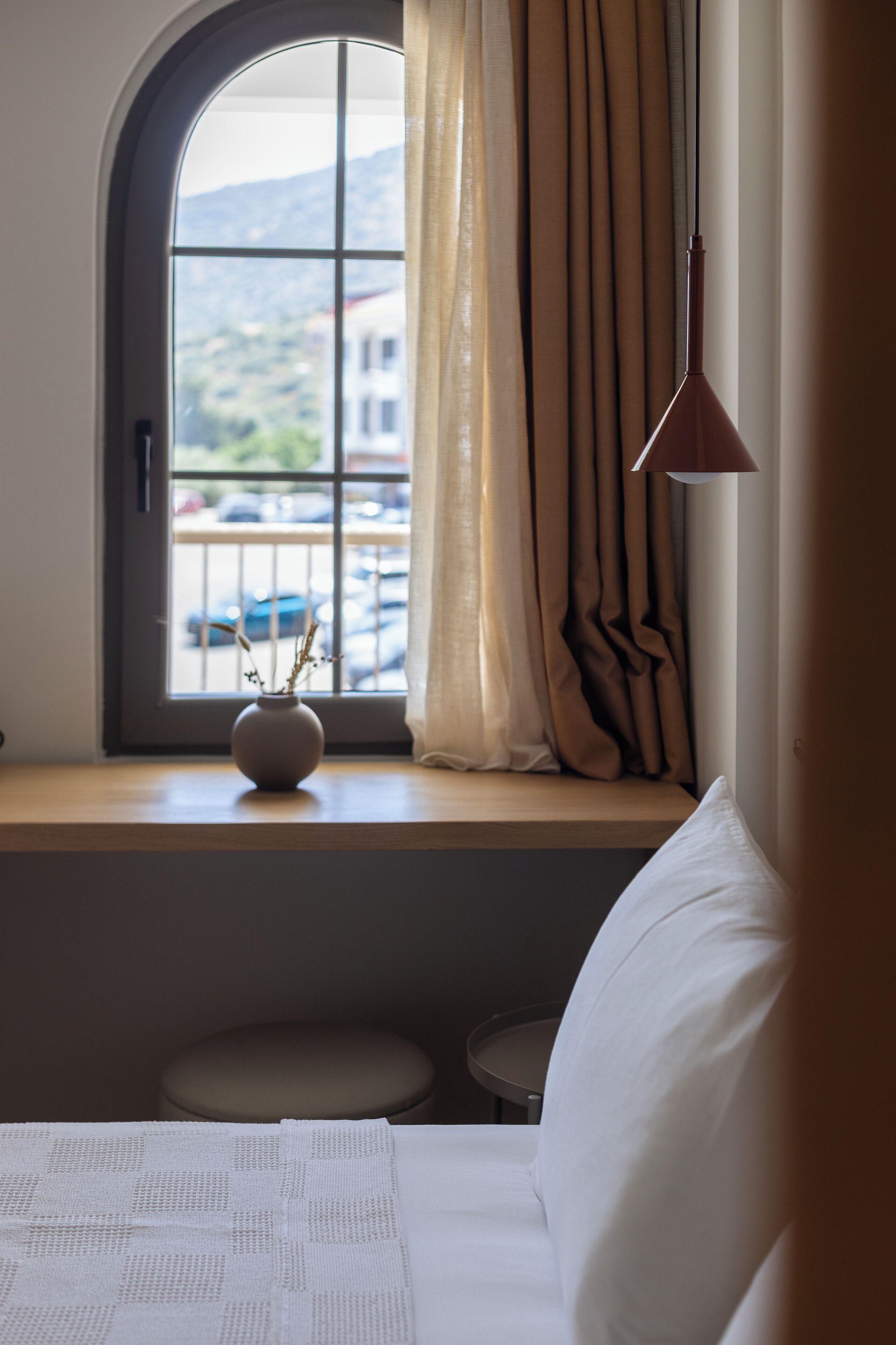 A room with a large arched window, a vase with branches on the sill, a bed with white bedding, a small round table, and a lamp.