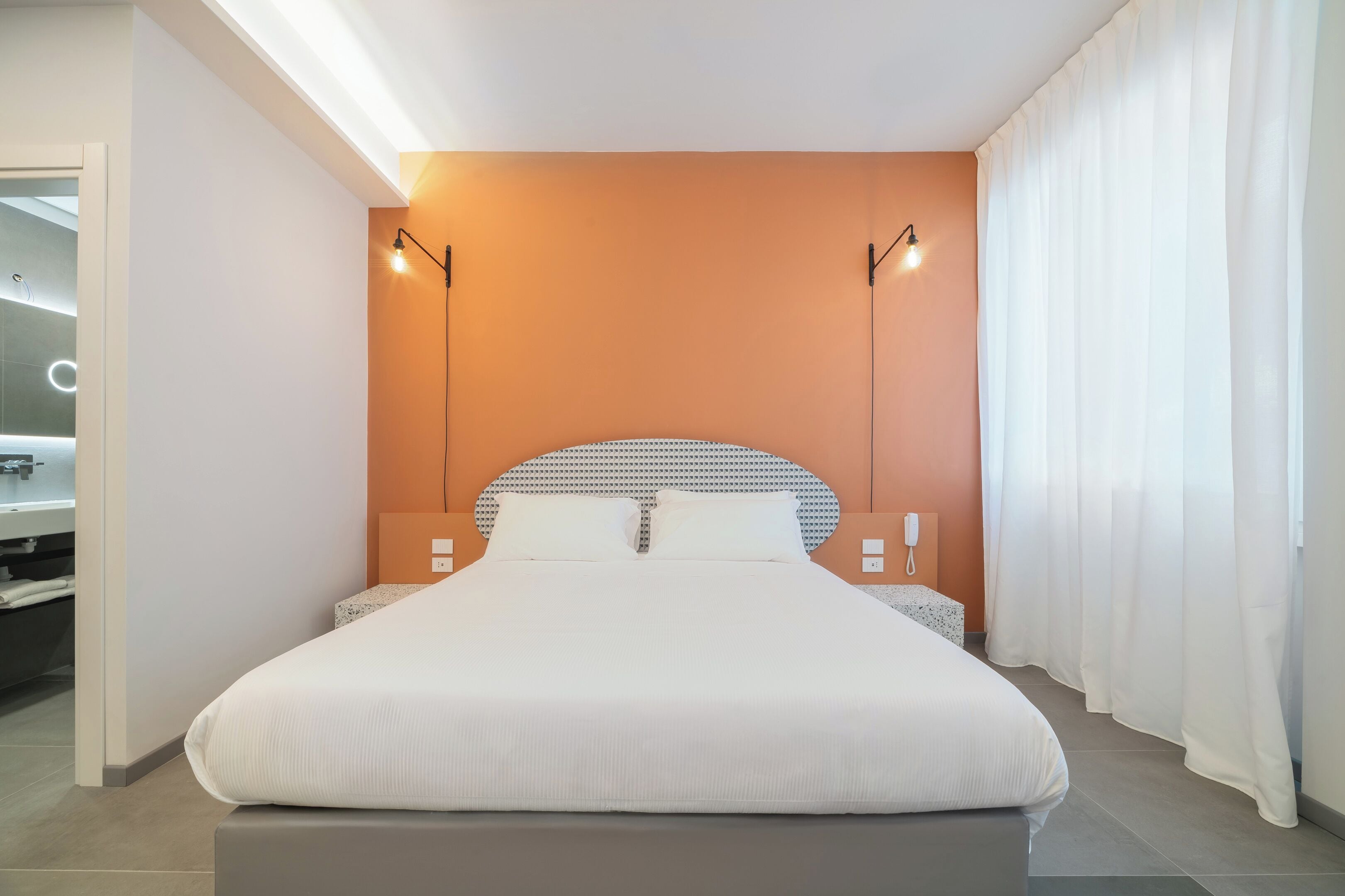 Una habitación de hotel moderna con una cama grande, dos mesitas de noche, una estantería empotrada y un baño a la vista.