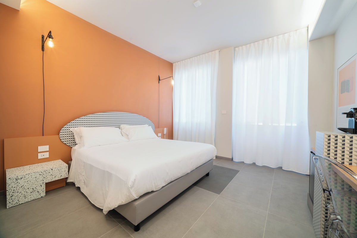 A modern bedroom with a large bed, a bedside table, a wall-mounted lamp, and a window with curtains.