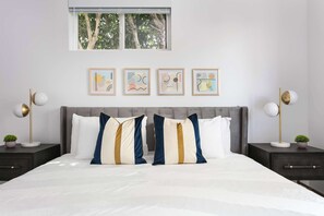 Une chambre à coucher comprenant un lit, deux tables de chevet, une fenêtre et quatre œuvres d’art encadrées accrochées au mur.