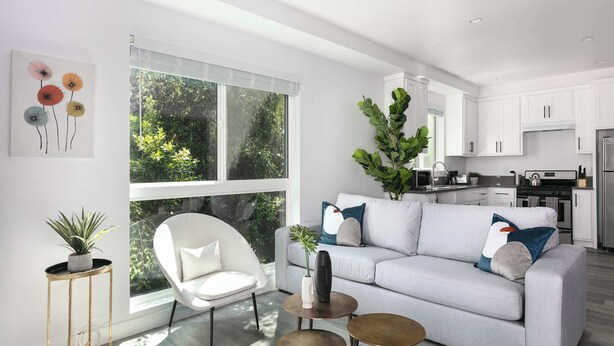 Een moderne woonkamer met een grijze bank, witte fauteuils en een eethoek, met een keuken op de achtergrond.
