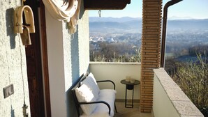 A balcony with a view of a valley and mountains, a white chair, a small round table, and a wooden door.