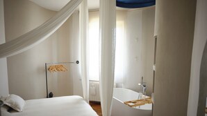 A bedroom with a large bed, a bedside table, a hanging light fixture, and a bathroom with a bathtub.