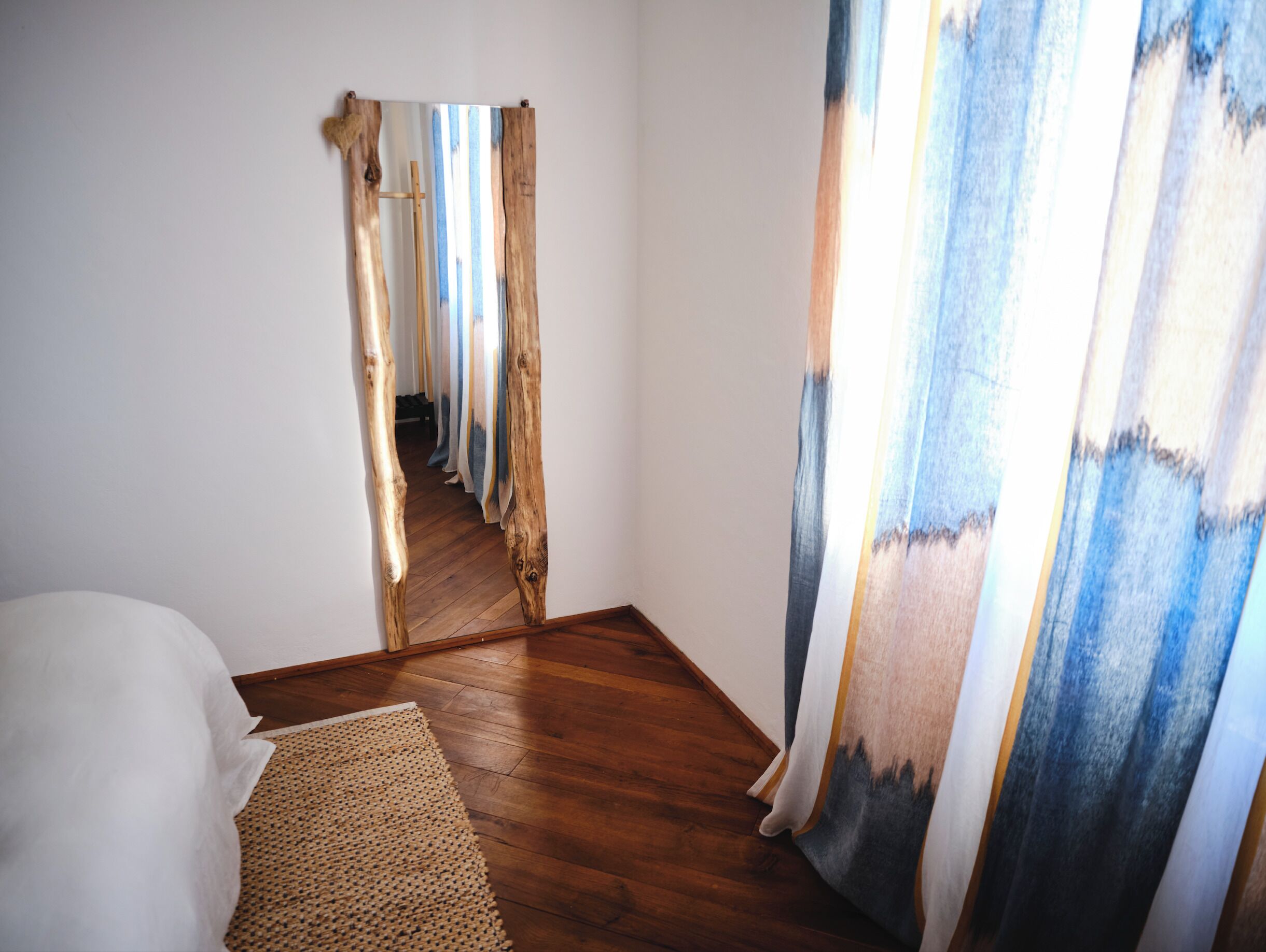A room with a wooden floor, a bed, and a view through a doorway to another room with curtains.