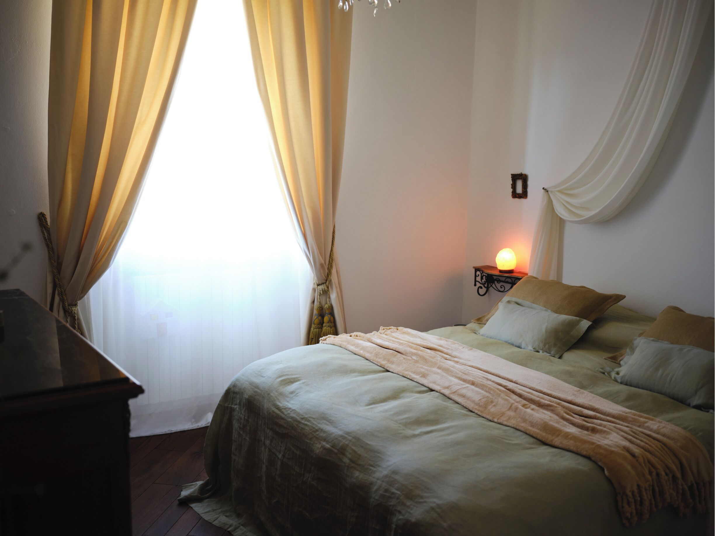 Un dormitorio con cama, cortinas, una lámpara de noche y un aplique en la pared.