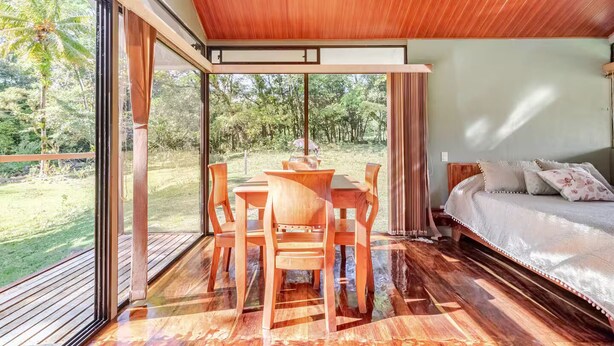 Una habitación amplia con una mesa y sillas de madera, una cama grande con almohadas y un ventanal corredero que da a un jardín exuberante.