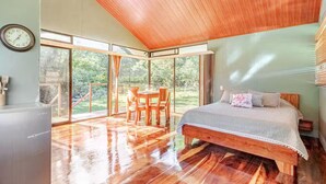 Een ruime slaapkamer met een houten plafond, een groot bed, een eettafel met stoelen en uitzicht op de omgeving door grote ramen.