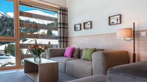 A cozy living room with a sofa, a coffee table with a vase of flowers, and a view of snow-capped mountains through large windows. - 3-room deluxe vacation apartment (Churwalden)