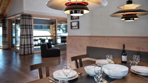 A dining area with a table set for four, including plates, glasses, and cutlery. There are two pendant lights above the table, and a bottle of wine is visible on the table. The room has wooden flooring and a brick wall in the background. - 3-room deluxe vacation apartment (Churwalden)