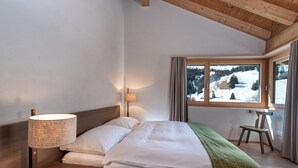 A bedroom with a bed, a nightstand, a lamp, a chair, and a window with a view of a snowy landscape. - 3-room deluxe vacation apartment (Churwalden)