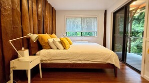 Een slaapkamer met een houten hoofdbord, een nachtkastje met een lamp, een bed met wit beddengoed en een raam met zonwering.