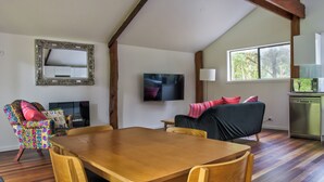 Ein Essbereich mit Holztisch und -stühlen, einem farbenfrohen Sessel, einer schwarzen Couch, einem an der Wand montierten Fernseher und einem Fenster mit Blick auf Bäume.
