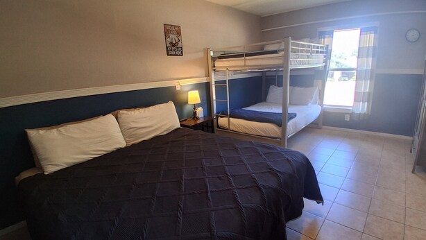 A hotel room with a large bed, a bunk bed, a bedside lamp, and a window with curtains.