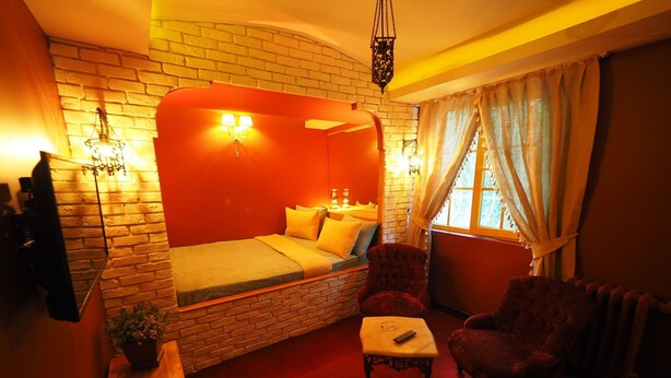 Une pièce chaleureuse avec un mur en briques, un lit avec une literie jaune, un fauteuil rouge, une petite table et une fenêtre avec des rideaux.