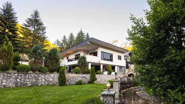 A modern two-story house with a stone wall, a small stream, and a patio area. - Villa Tomi Nature Retreat (Sarajevo)