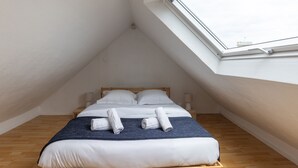 A bedroom with a skylight, wooden flooring, a bed with white linens, and two rolled towels on the bed.