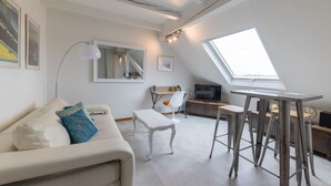 A modern loft apartment with a high ceiling, a skylight, a white sofa, a small dining table, and a TV.