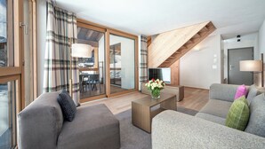 En moderne stue med sofa, skammel og sofabord. Der er en trappe med træbjælker og udsigt til naturen gennem store vinduer.