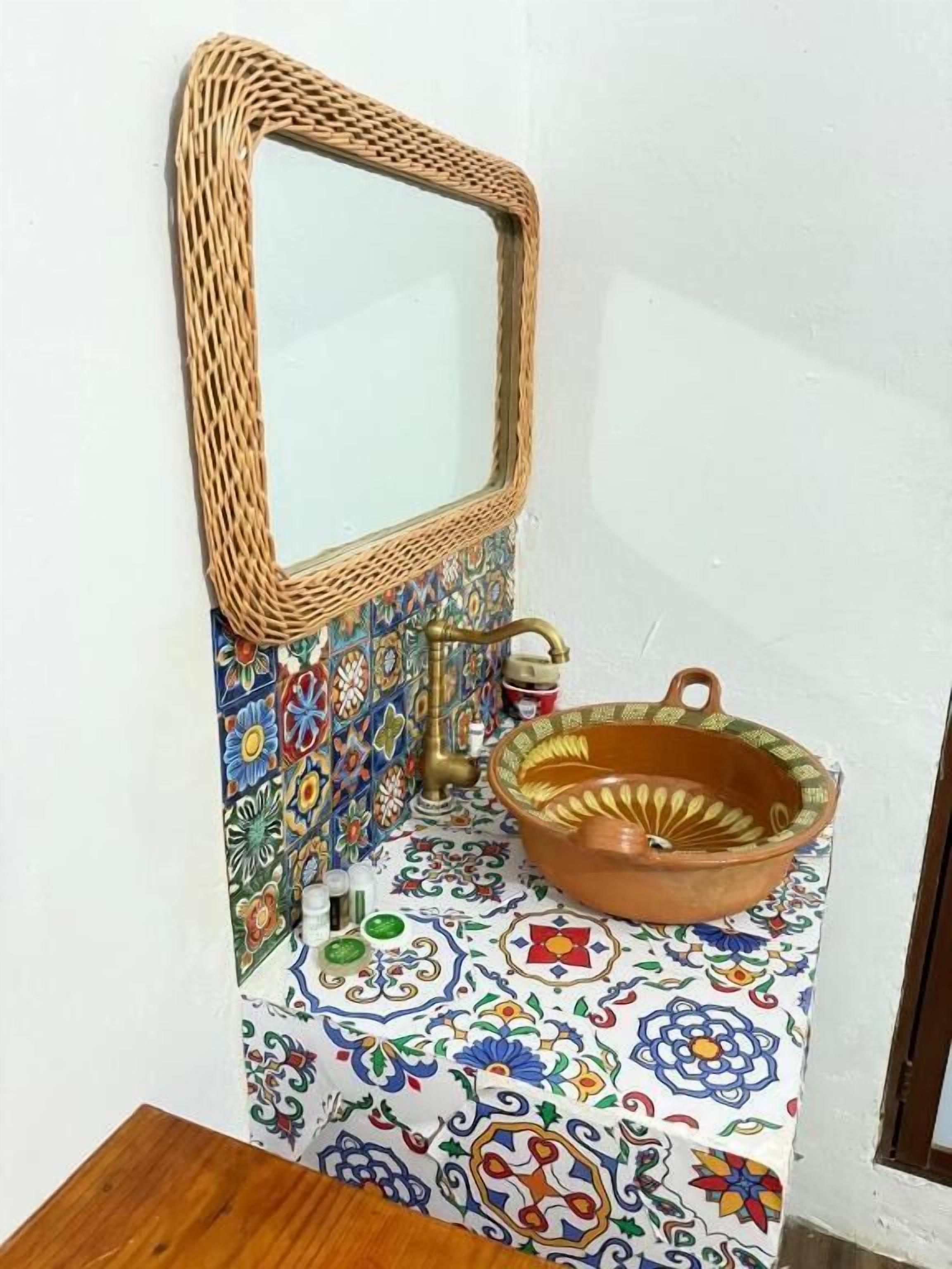 Une salle de bain avec un miroir à motifs tissés, un lavabo en terre cuite et un mur recouvert de carreaux aux motifs floraux.