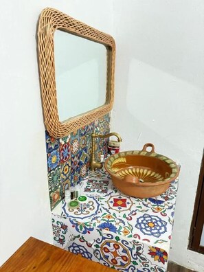 A bathroom with a woven mirror, a terracotta basin, and a tiled wall with floral patterns.