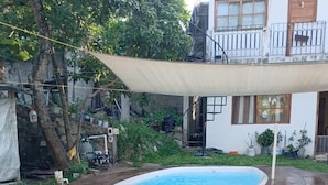 A backyard with a swimming pool, wooden steps, a covered area, and a house in the background.