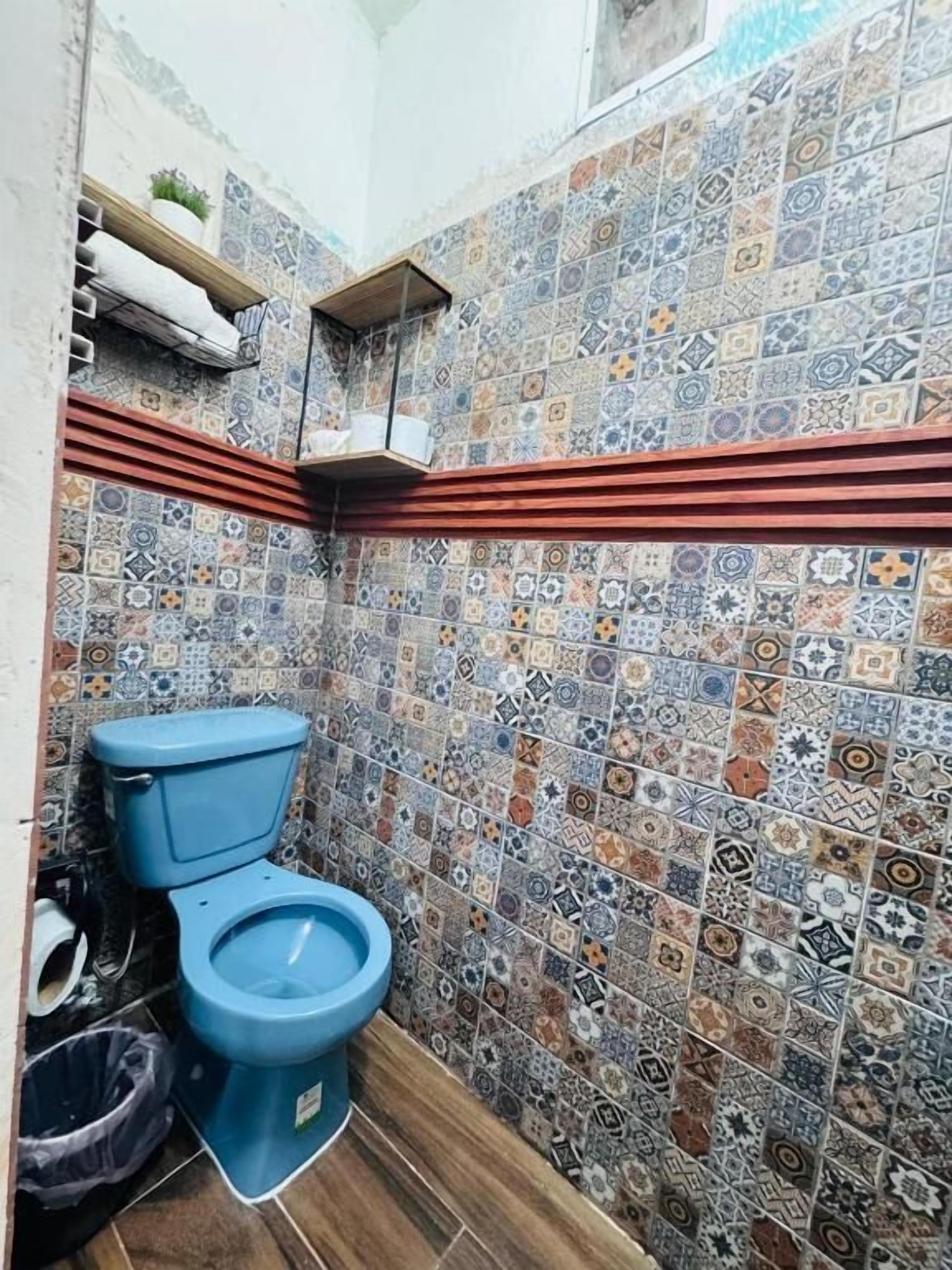 Un baño con inodoro azul, baldosas estampadas y suelo de madera.