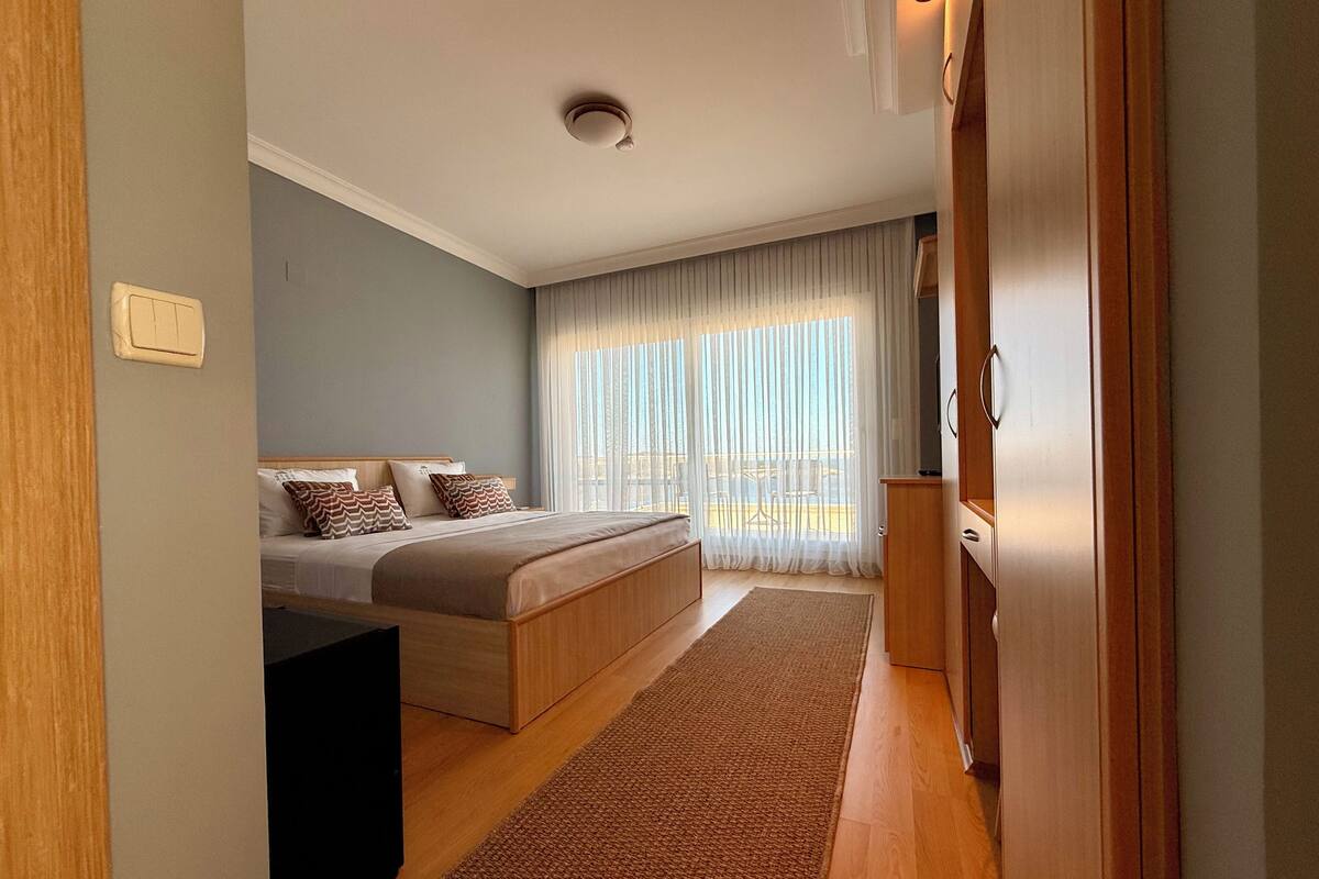 Une chambre à coucher avec un lit, une armoire en bois, un bureau et une grande fenêtre ornée de rideaux légers.