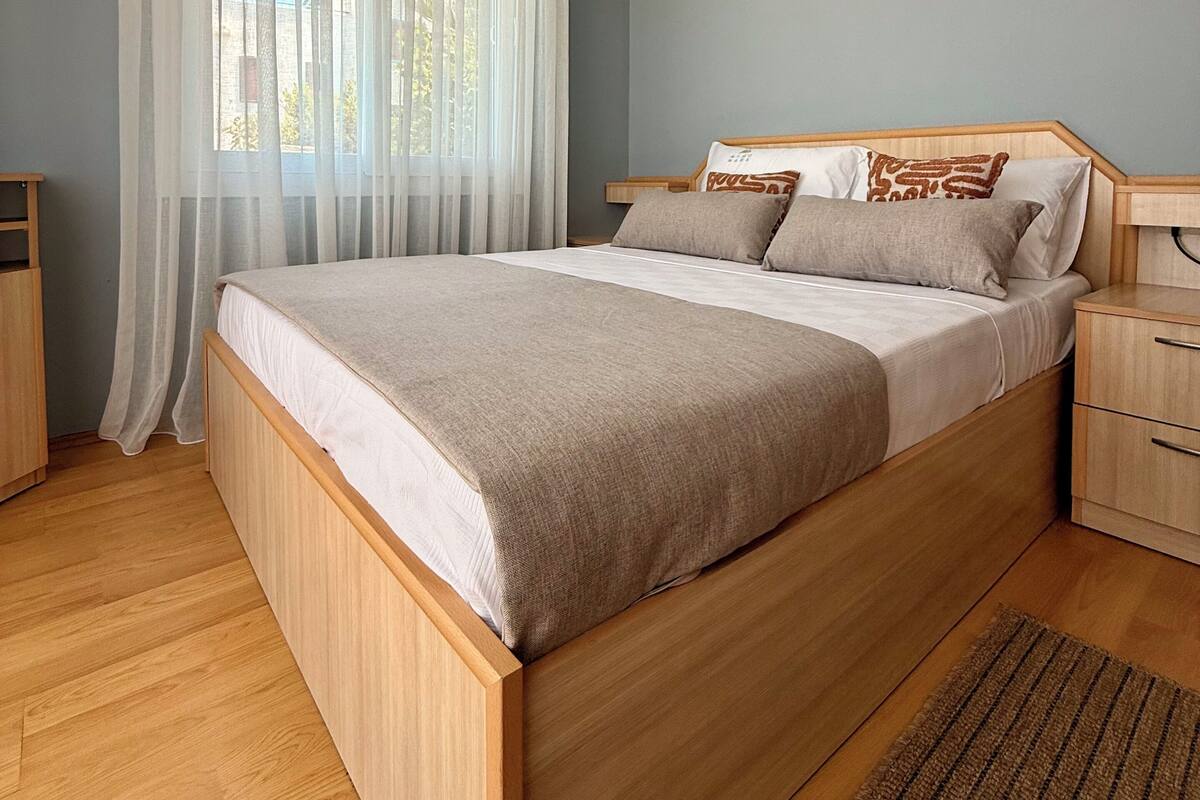Une chambre à coucher avec un lit en bois, une table de chevet, une commode et une fenêtre ornée de rideaux légers.