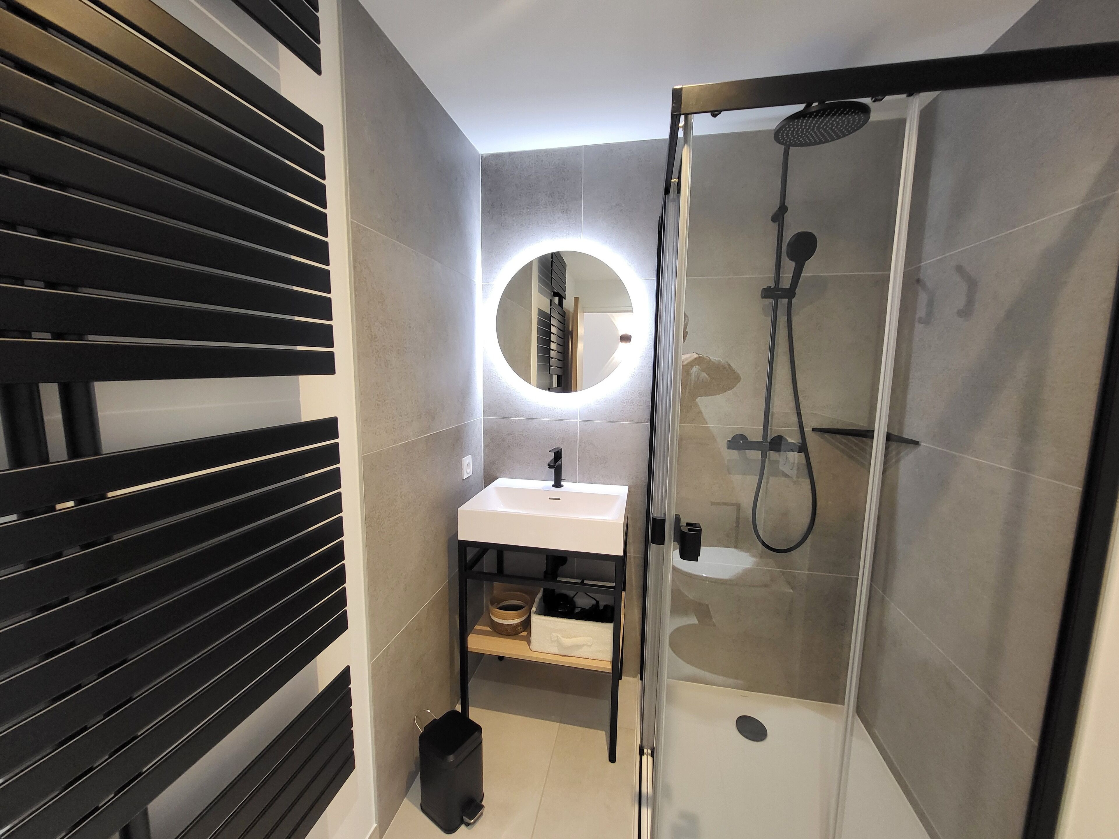 A modern bathroom with a glass-enclosed shower, a round mirror with a light, and a black-framed sink.