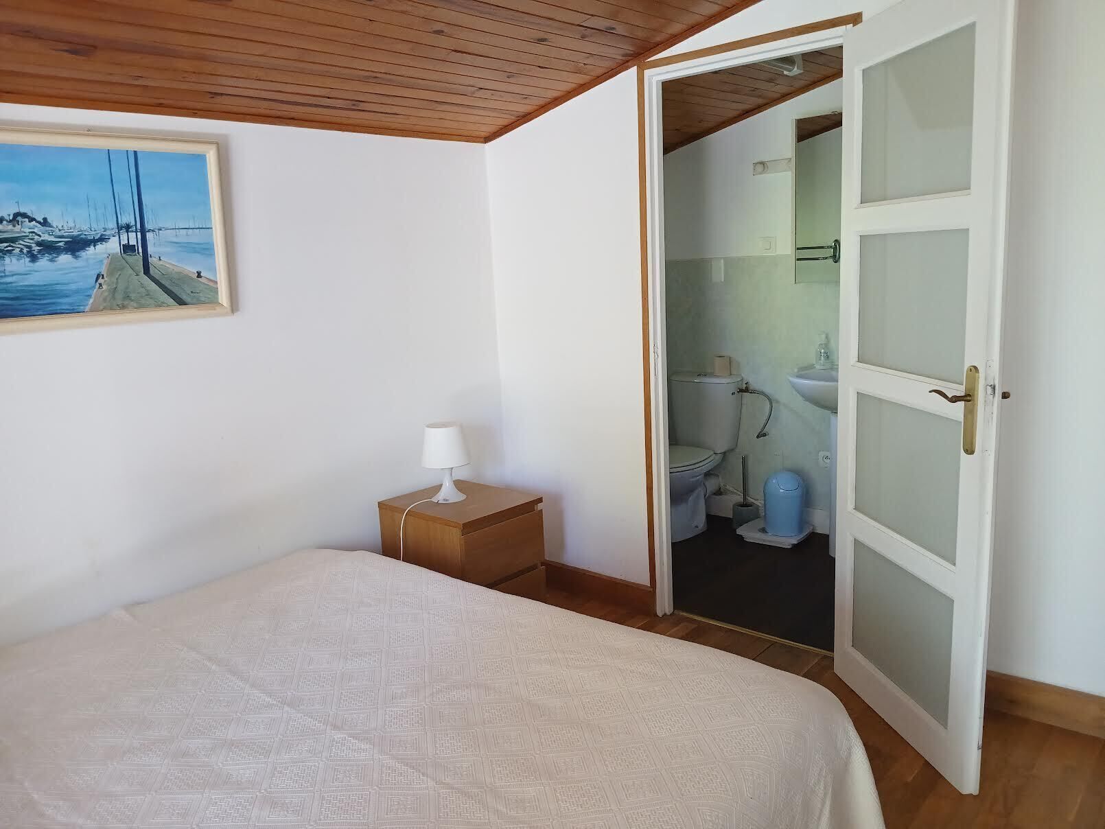 Un dormitorio con una cama, una mesita de noche con una lámpara, un cuadro de un puerto deportivo y una puerta abierta que da a un baño.