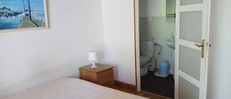 Un dormitorio con una cama, una mesita de noche con una lámpara, un cuadro de un puerto deportivo y una puerta abierta que da a un baño.