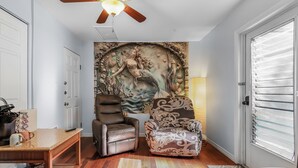 Un salon avec un ventilateur de plafond, une fresque murale, un fauteuil en cuir marron, un fauteuil à motifs et une table en bois.