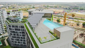 Un edificio moderno con piscina sul tetto, circondato da vegetazione e adiacente ad altri edifici.