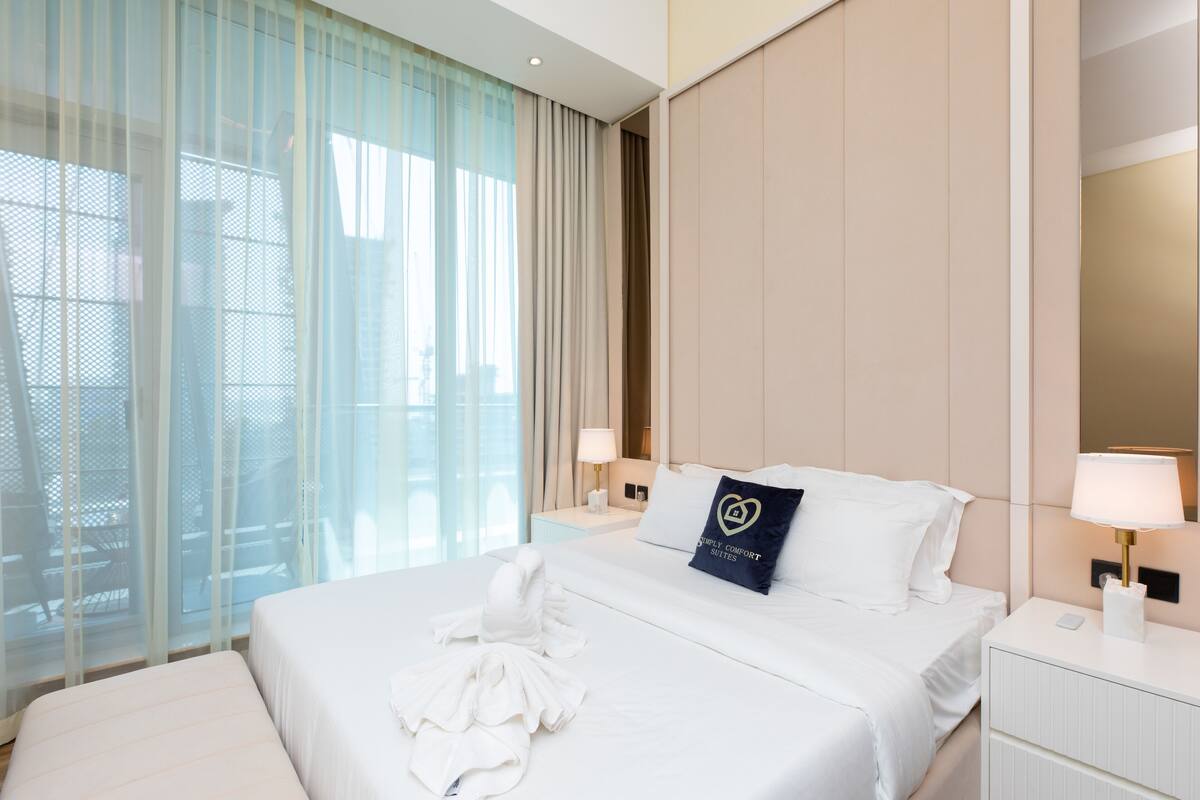 A hotel room with a large bed, a bedside table with a lamp, a window with sheer curtains, and a view of the outdoors.