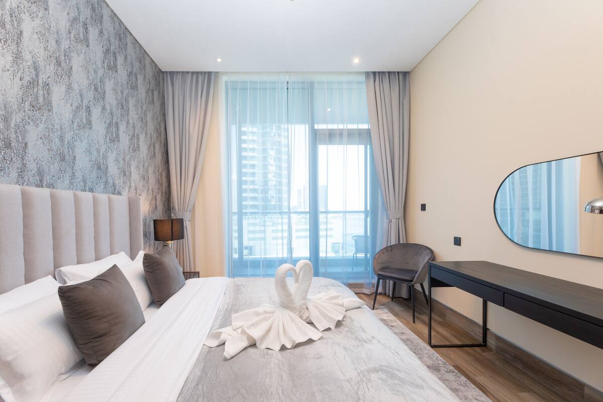 A modern hotel room with a large bed, a desk, and a view of the city through large windows.