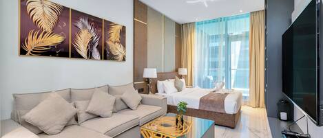 A modern hotel room with a sofa, a bed, a flat-screen TV, and a large window with curtains.