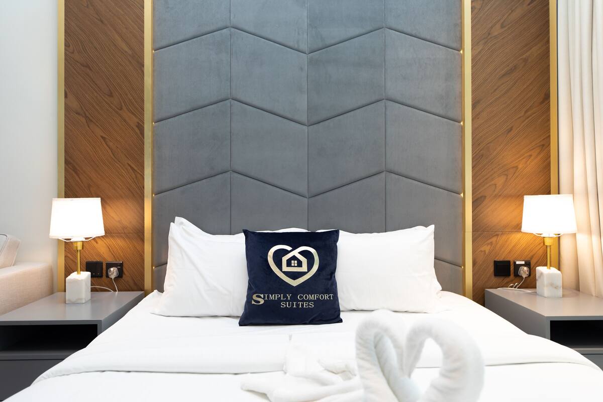 A hotel room with a large bed, two bedside tables with lamps, a heart-shaped pillow with a logo, and a towel on the bed.