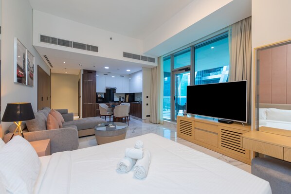 A modern hotel room with a large bed, a flat-screen TV, a sofa, a dining area, and a view of a building through the window.