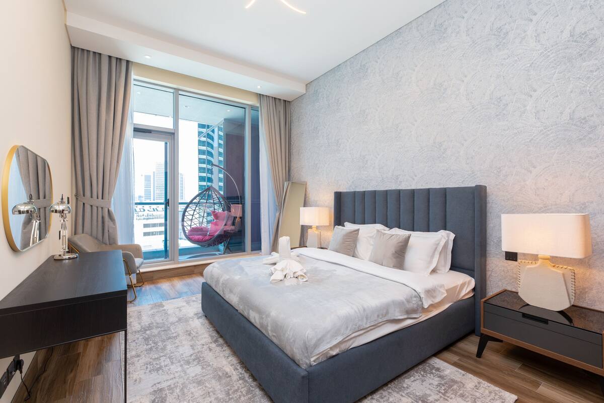 A modern hotel room with a large bed, a desk, a chair, and a balcony with a view of the city.