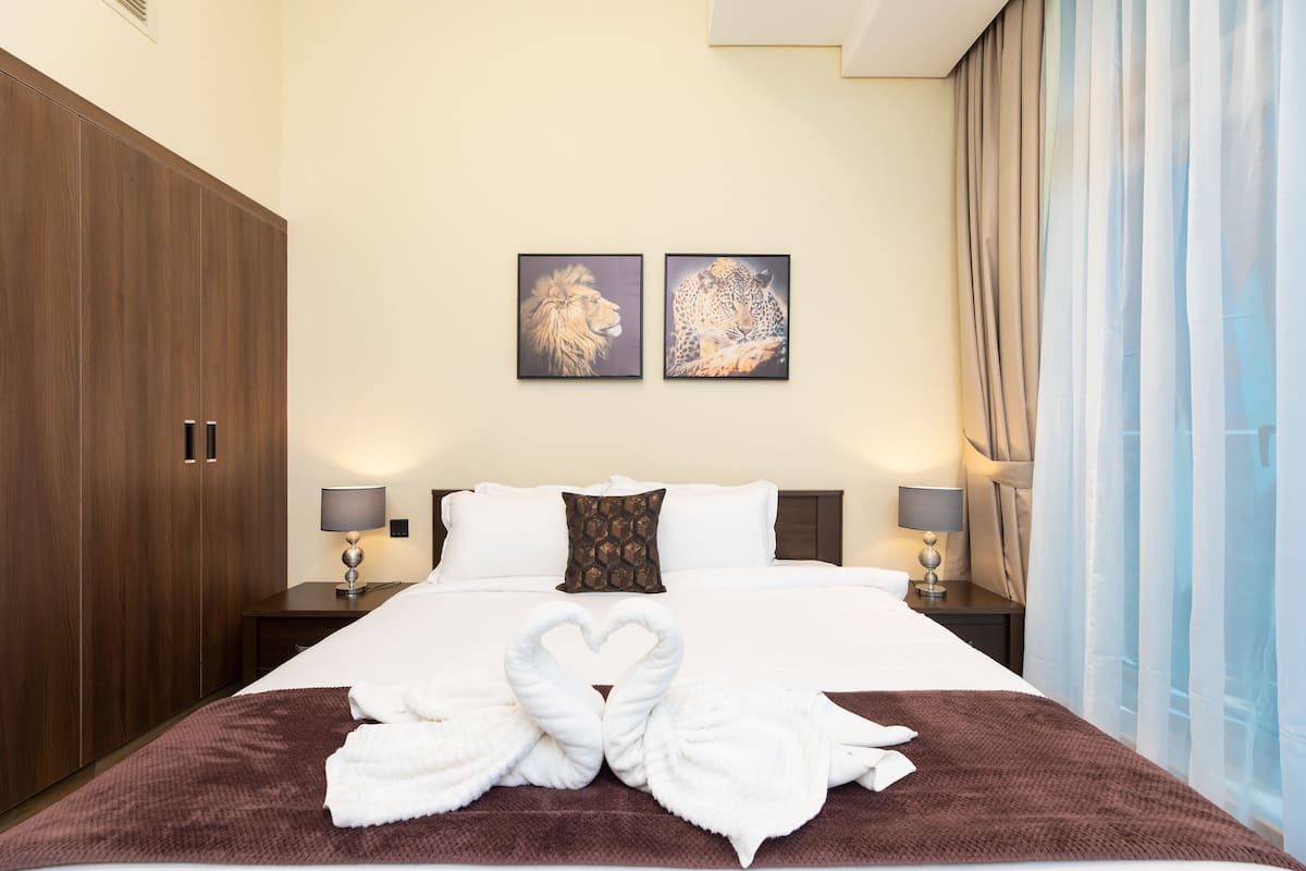 A hotel room with a large bed, two wall-mounted paintings, a wardrobe, and a nightstand with a lamp.