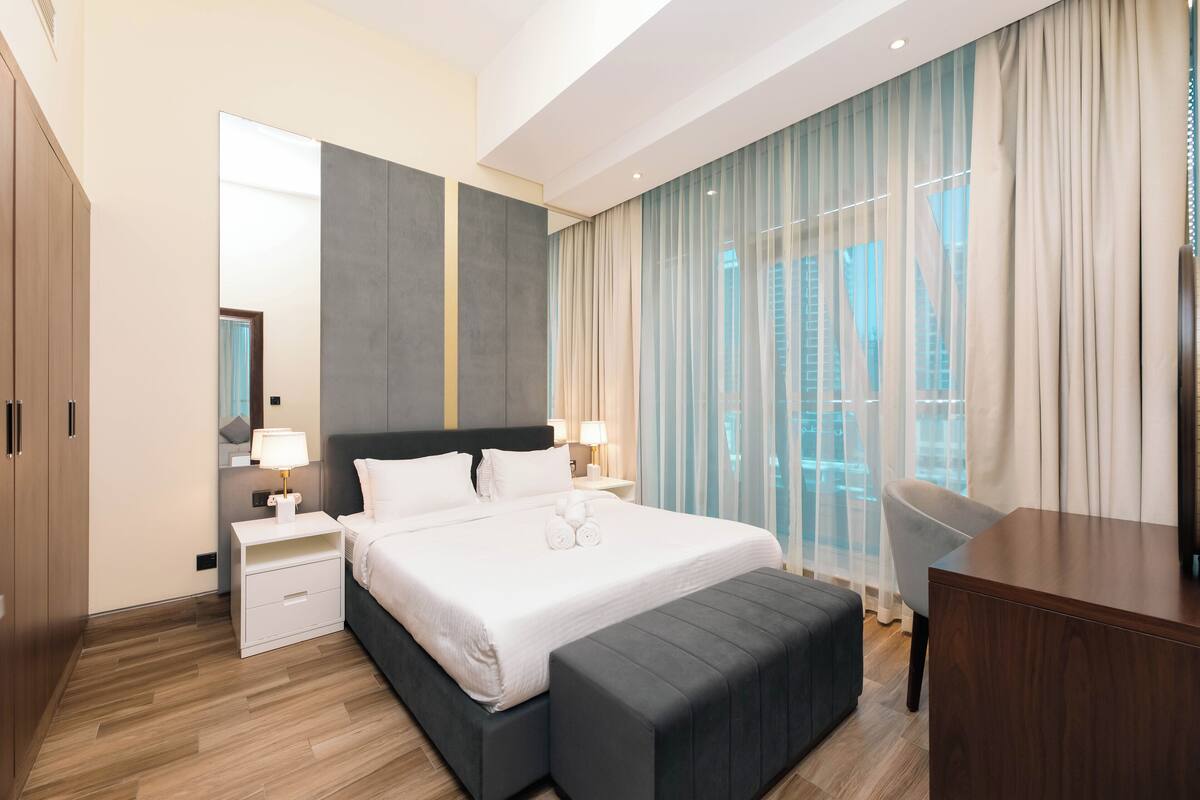 A modern hotel room with a large bed, bedside tables, a desk, and a view of the city through large windows.