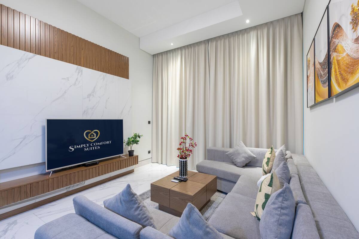 A modern living room with a flat-screen TV, a sofa set, a coffee table, and a large window with curtains.