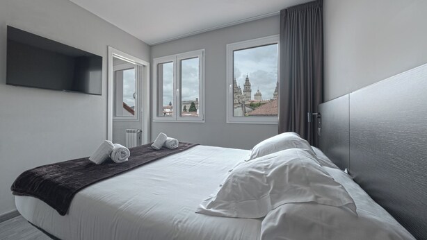 Una habitación de hotel con una cama, televisor de pantalla plana, tres ventanas y vista a un paisaje urbano.
