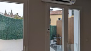 Habitación con puerta corrediza de vidrio que da a un balcón, aire acondicionado en el techo y vista a un edificio con tejado de tejas.