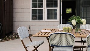 A patio with a wooden table and chairs, a vase with flowers, and a house with a garage door. - Amazing 3-bedroom cottage. 5 Min-walk to beach. Beach side of Oak Island Drive (Oak Island)