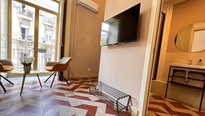 A hotel room with a large window, a flat-screen TV mounted on the wall, a small round table with chairs, a bench, and a tiled floor with a geometric pattern. - Dimore del Plebiscito B&B (Catania)
