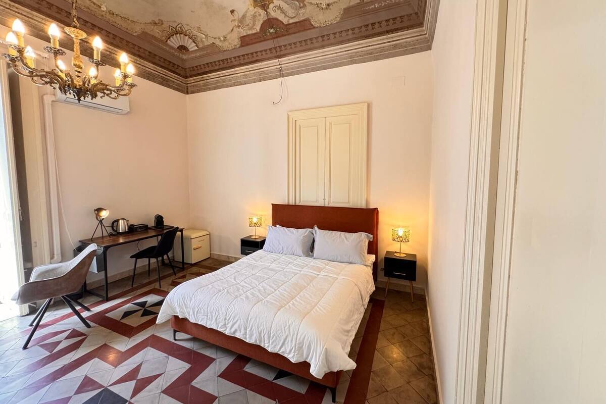 A bedroom with a large bed, a desk, and a chair. There is a chandelier above the bed and a lamp on the desk.